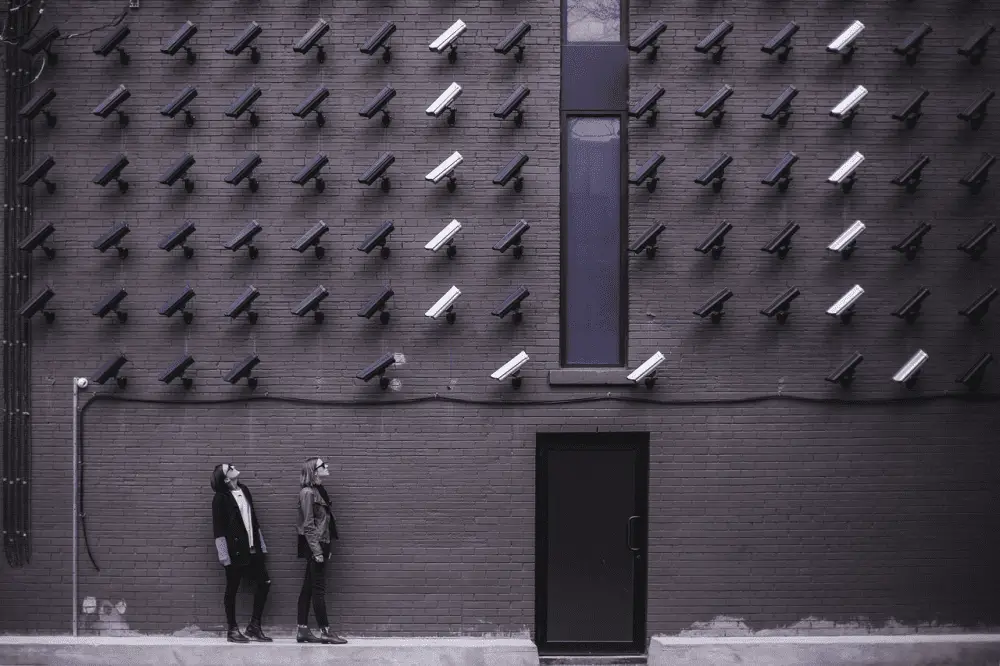 Two individuals positioned near a building's brick wall under surveillance cameras.