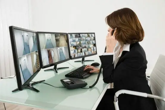 Young Female Operator Looking At Multiple Camera Footage On Computers While Talking On Phone